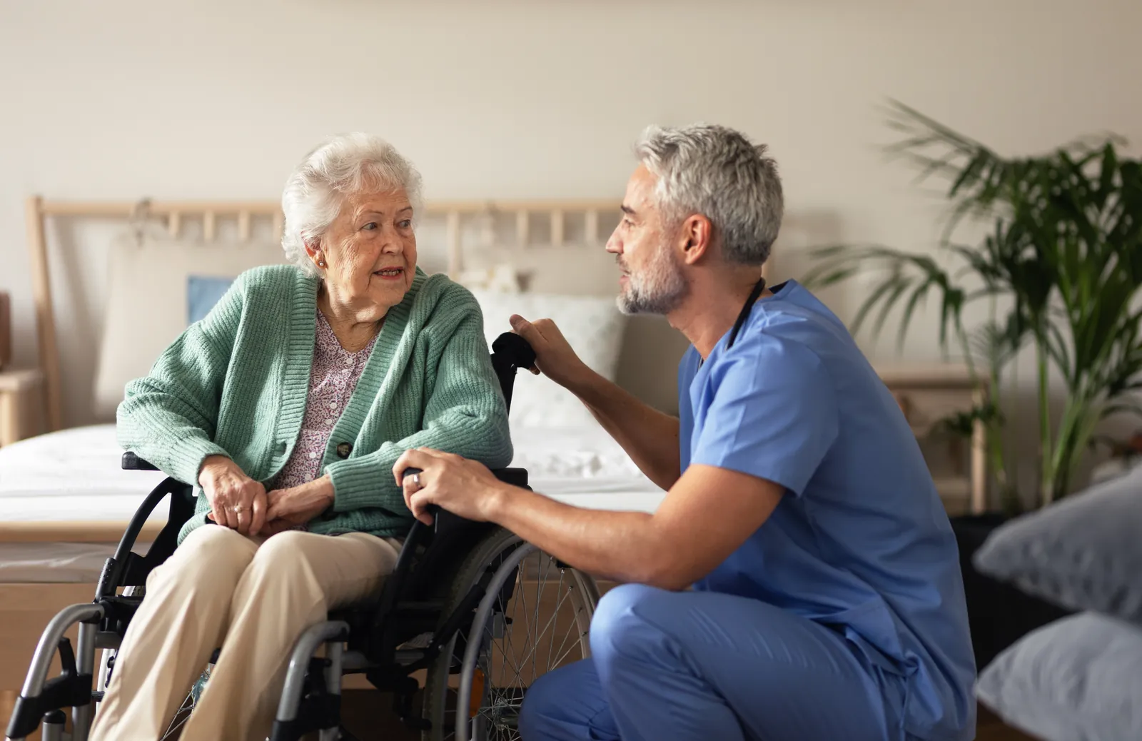 Carer crouching beside an older man's wheelchair in a bedroom, mid-conversation